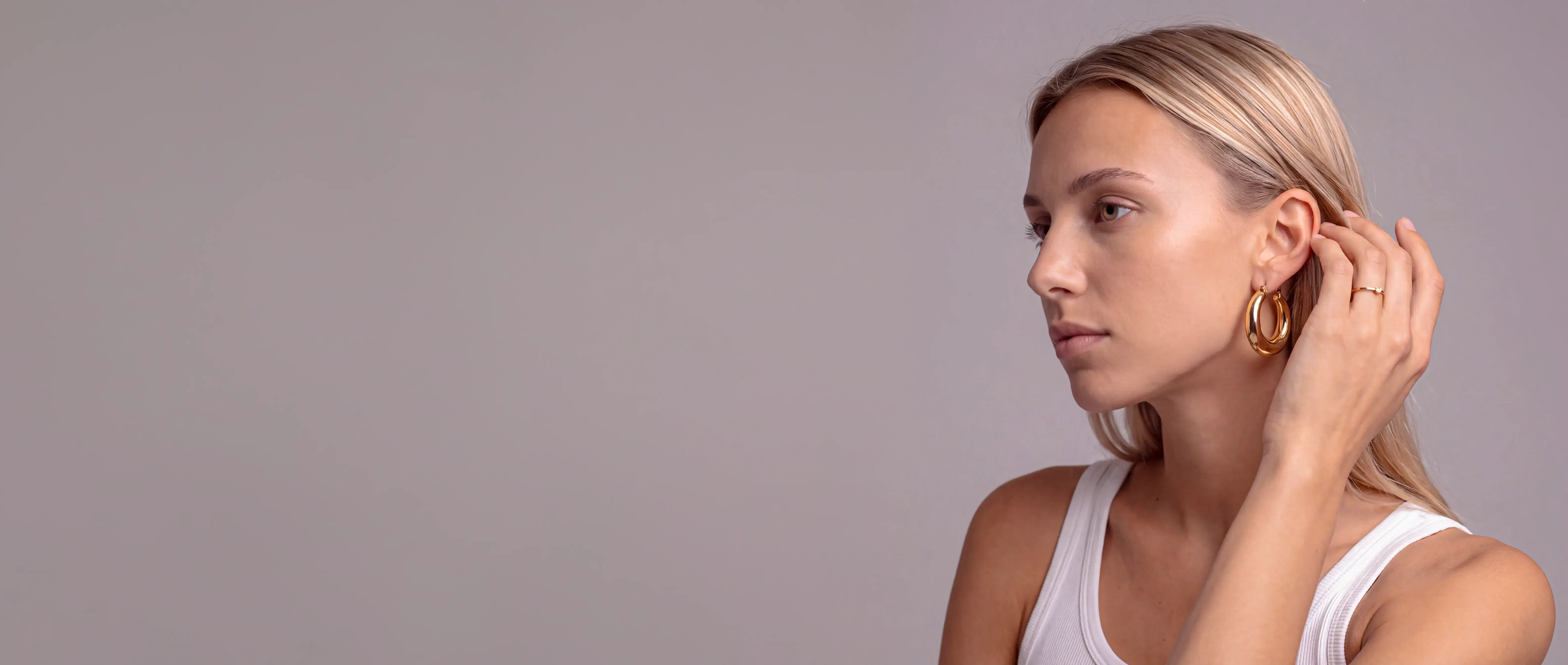 A model touching a gold hoop earring, in profile against a warm grey studio — Epox campaign still