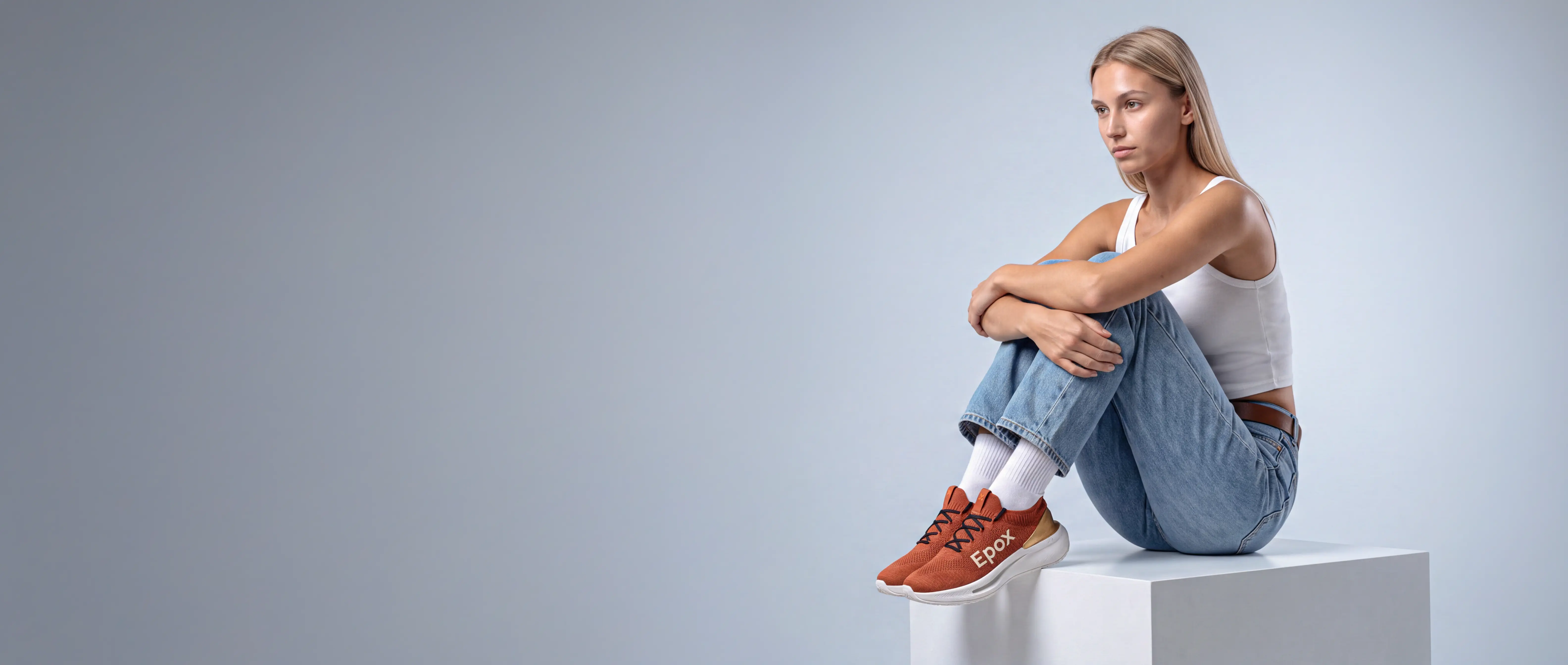 A model seated on a white plinth in rust knit sneakers — Epox campaign still